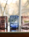 Four colorful glass tumblers on a reflective surface with a blurred natural background