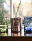 Four colorful glass tumblers on a reflective surface with a blurred natural background
