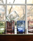 Set of colorful glass tumblers on a reflective surface with a window background