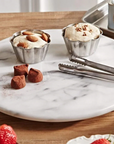 Two metal dessert cups with ice cream on a marble platter with truffles and tongs.