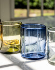 Four glass tumblers of different colors on a reflective surface with a blurred outdoor background