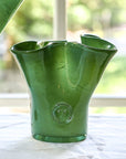A handcrafted green glass vase with a lettuces leaf design on a table, with a blurred background.