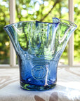 A handcrafted blue glass vase with a lettuce leaf pattern, placed on a table with a blurred background of greenery.