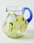 Clear glass pitcher with blue handle filled with lemon and lime slices infused drink and ice on a white background