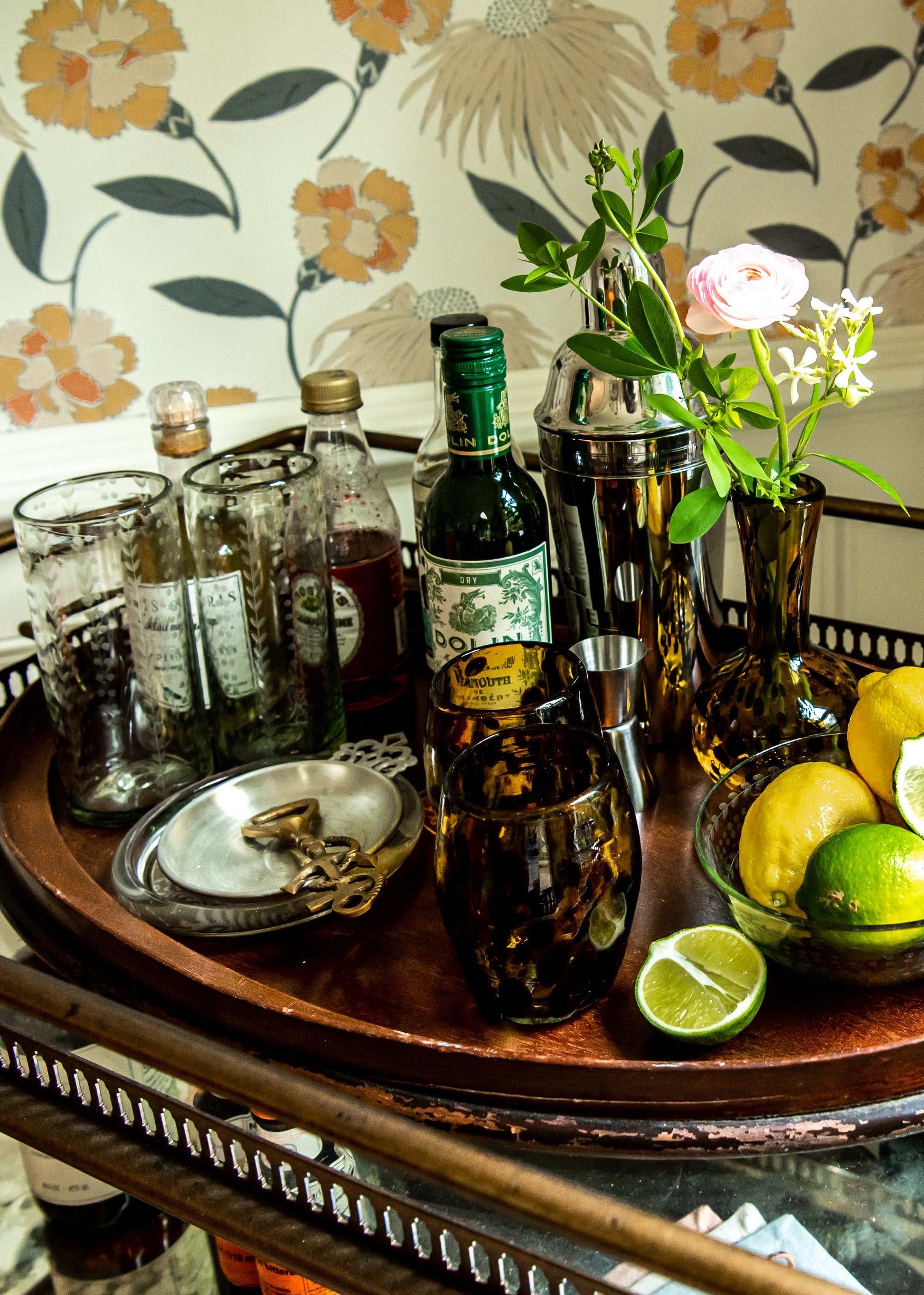 Bar tray with bottles, glasses, and a floral arrangement against a patterned wall.