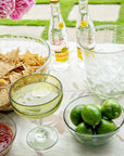 Outdoor setting with tortilla chips on a clear etched bowl, limes in a small etched bowl, a glass of limeade, and bottles on a table.