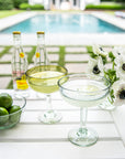 Outdoor setting with margarita glasses filled with cocktails, limes, and flowers by a poolside.