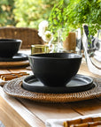 Dining table setting with black bowls, plates, and glasses on a wooden table outdoors.