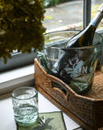 A clear glass champagne bucket with hand-etched fern frond motif, containing a champagne bottle, placed on a table with a blue and white floral tablecloth.