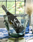 A clear glass champagne bucket with hand-etched fern frond motif, containing a champagne bottle, placed on a table with a blue and white floral tablecloth.