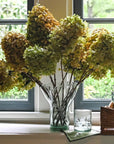 Bouquet of hydrangeas in a vase on a windowsill with a glass and basket in the background.