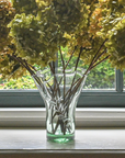 A clear glass vase with a fern leaf motif, containing stems and placed on a windowsill with hydrangea flowers in the background.