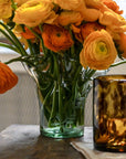 A clear glass vase with a fern motif, containing orange and yellow flowers, placed on a surface with a brown glass object beside it.