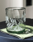 A clear rocks glass with hand-etched fern frond motif, placed on a green coaster on a dark surface.