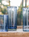 Three blue glass tumblers on a woven surface with a blurred natural background