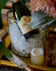 Bottle of champagne in a glass with ice next to a glass of champagne on a wooden tray.