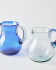 Two glass pitchers, one blue with a clear handle and one clear with a blue handle, on a white background