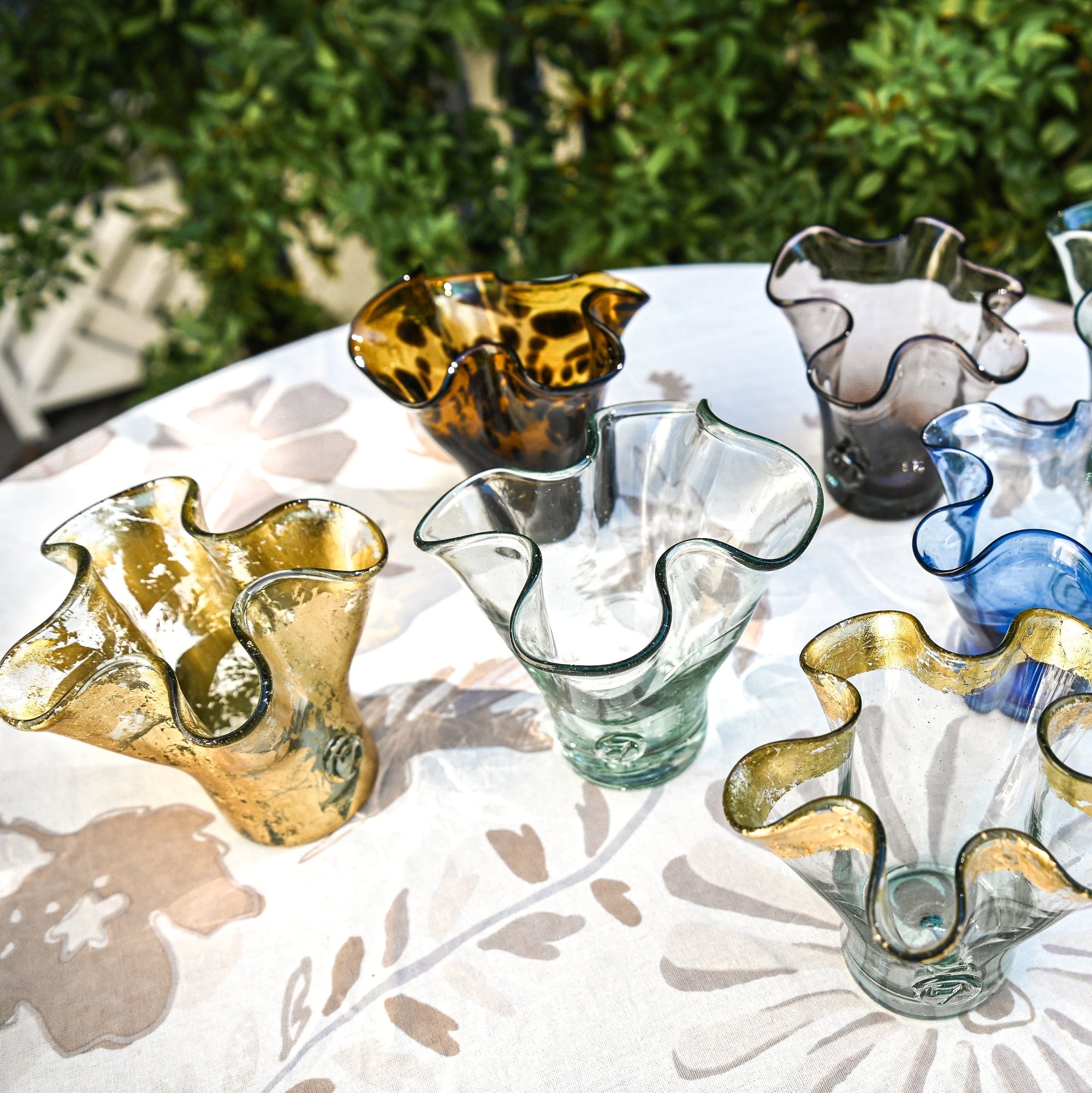 A handcrafted colored glass vase with a lettuce leaf design, made from recycled glass, displayed on a table with a window in the background.
