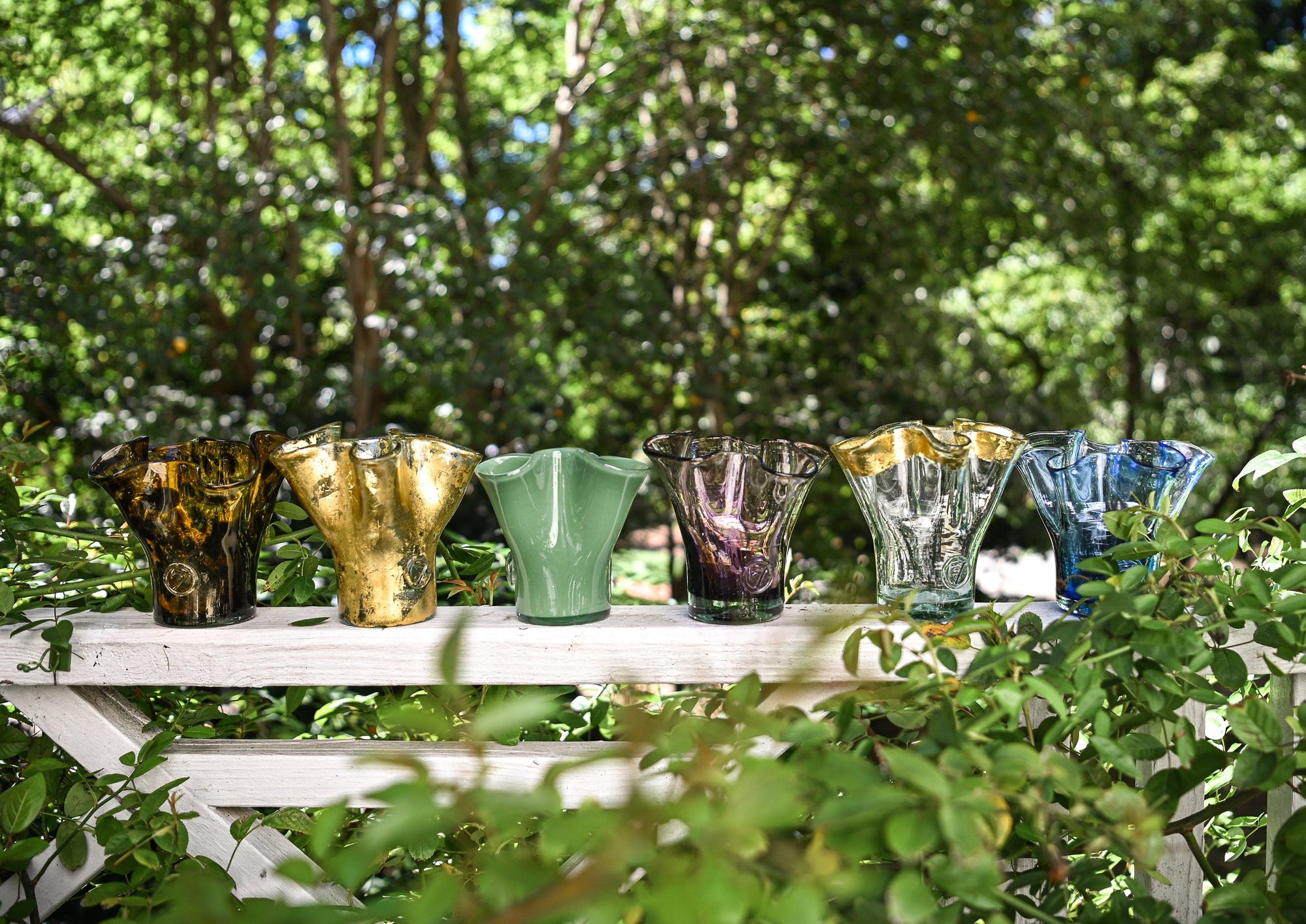 Colorful glass vases on a white table outdoors with greenery in the background