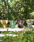 Colorful glass vases on a white table outdoors with greenery in the background