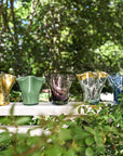 Colorful glass vases on a white table outdoors with greenery in the background