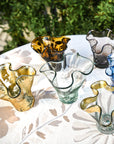 A handcrafted colored glass vase with a lettuce leaf design, made from recycled glass, displayed on a table with a window in the background.
