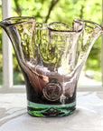 A handcrafted smokey amethyst colored glass vase with a lettuce leaf design, made from recycled glass, displayed on a table with a window in the background.