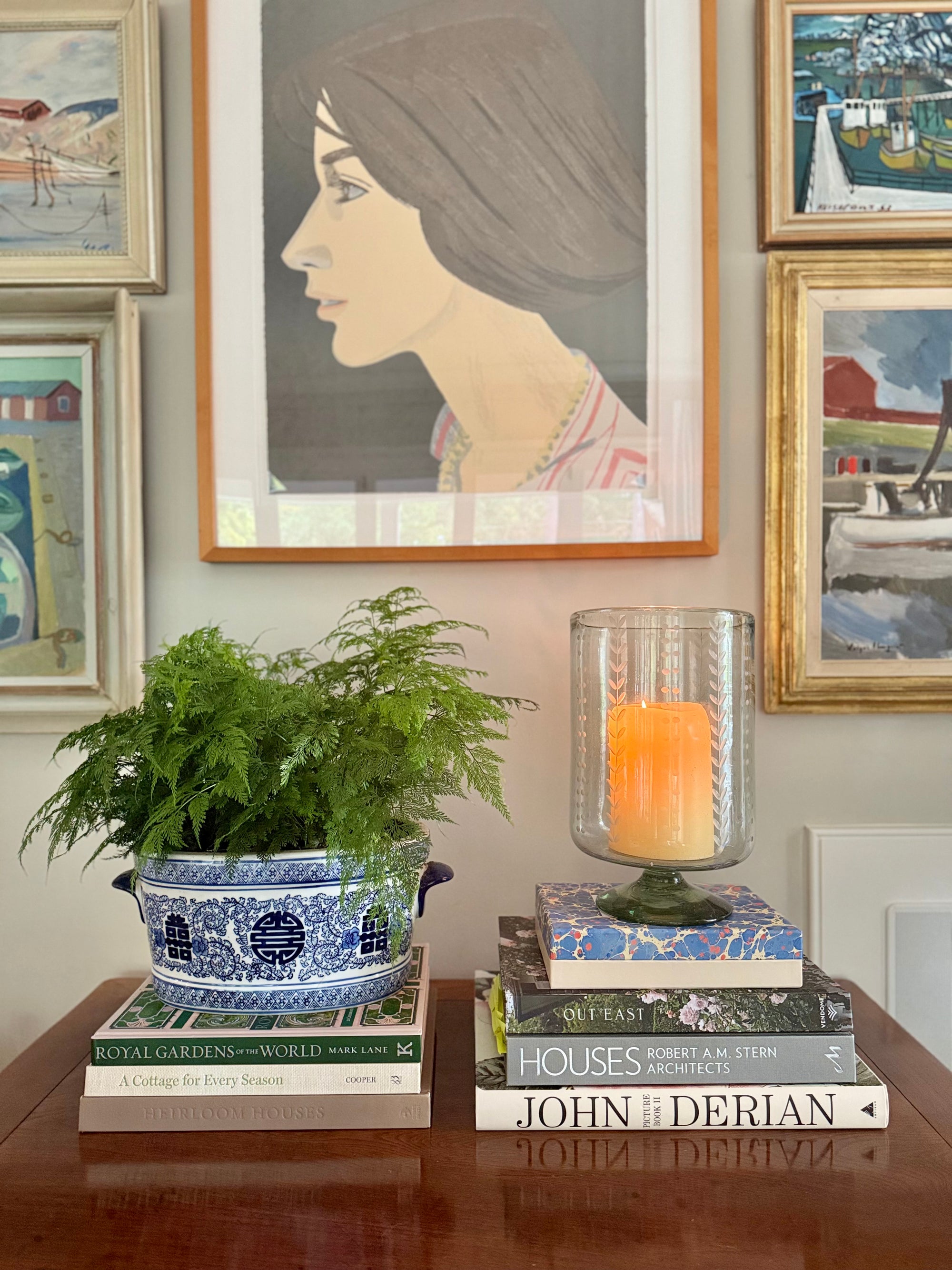 Decorative setup with books, a candle inside a recycled glass candleholder, and a potted plant against a wall with framed art.