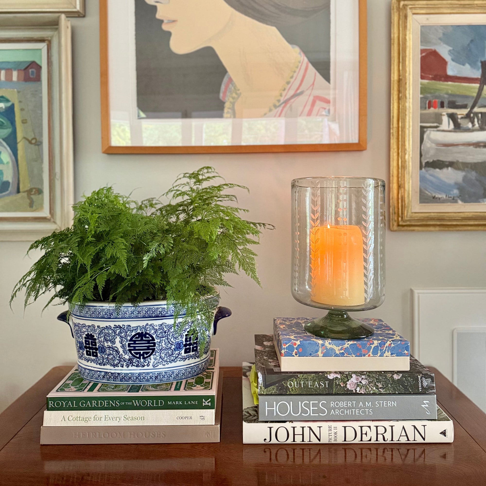 Decorative setup with books, a candle inside a recycled glass candleholder, and a potted plant against a wall with framed art.