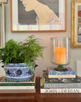Decorative setup with books, a candle inside a recycled glass candleholder, and a potted plant against a wall with framed art.
