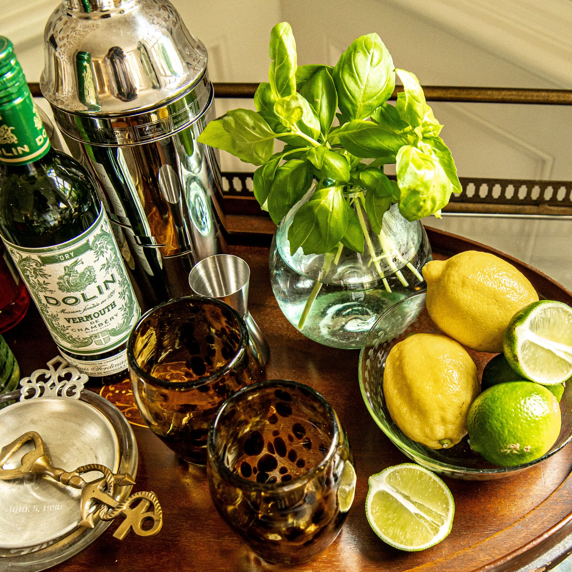 Bar setup with Dolin Vermouth bottle, cocktail shaker, glasses, lemons, limes, and basil on a wooden tray.