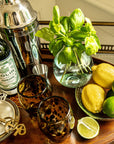 Bar setup with Dolin Vermouth bottle, cocktail shaker, glasses, lemons, limes, and basil on a wooden tray.