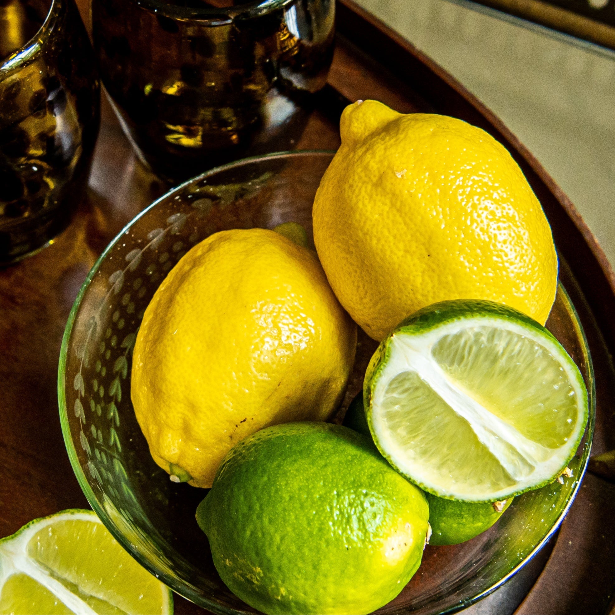 Bowl of lemons and limes on a wooden tray with glasses in the background.