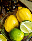 Bowl of lemons and limes on a wooden tray with glasses in the background.