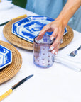 A hand placing down an iridescent purple rocks glass with a hand-etched heart design on a table set with blue and white dishes and cutlery.