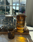 Decorative ceramic teapot, amber glass pitcher, and tumblers on a tray with a window view in the background.