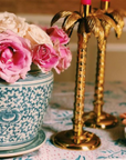 Gold palm tree candle holders with pink candles, a vase of pink roses, and a stack of plates on a table.