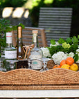 A scalloped rattan tray with a thick glass insert, displayed outdoors, used to serve beverages and snacks.