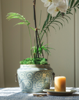 Decorative pot with plants and a candle on a table against a neutral wall.