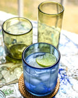 Blue glass with lime wedge on a woven coaster, surrounded by green glasses on a decorative tray.