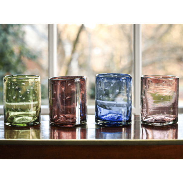 A set of four colored glass rocks glasses etched with stars, placed on a table with a window in the background.