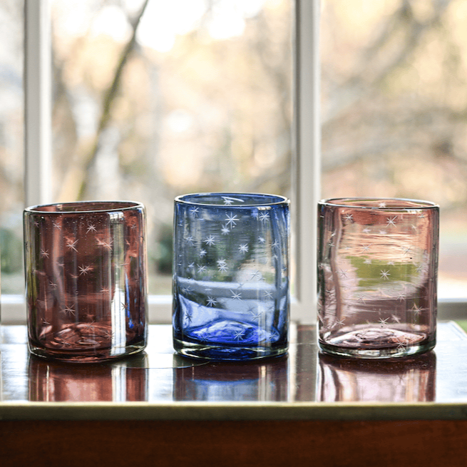 Four colorful glass tumblers on a reflective surface with a blurred natural background