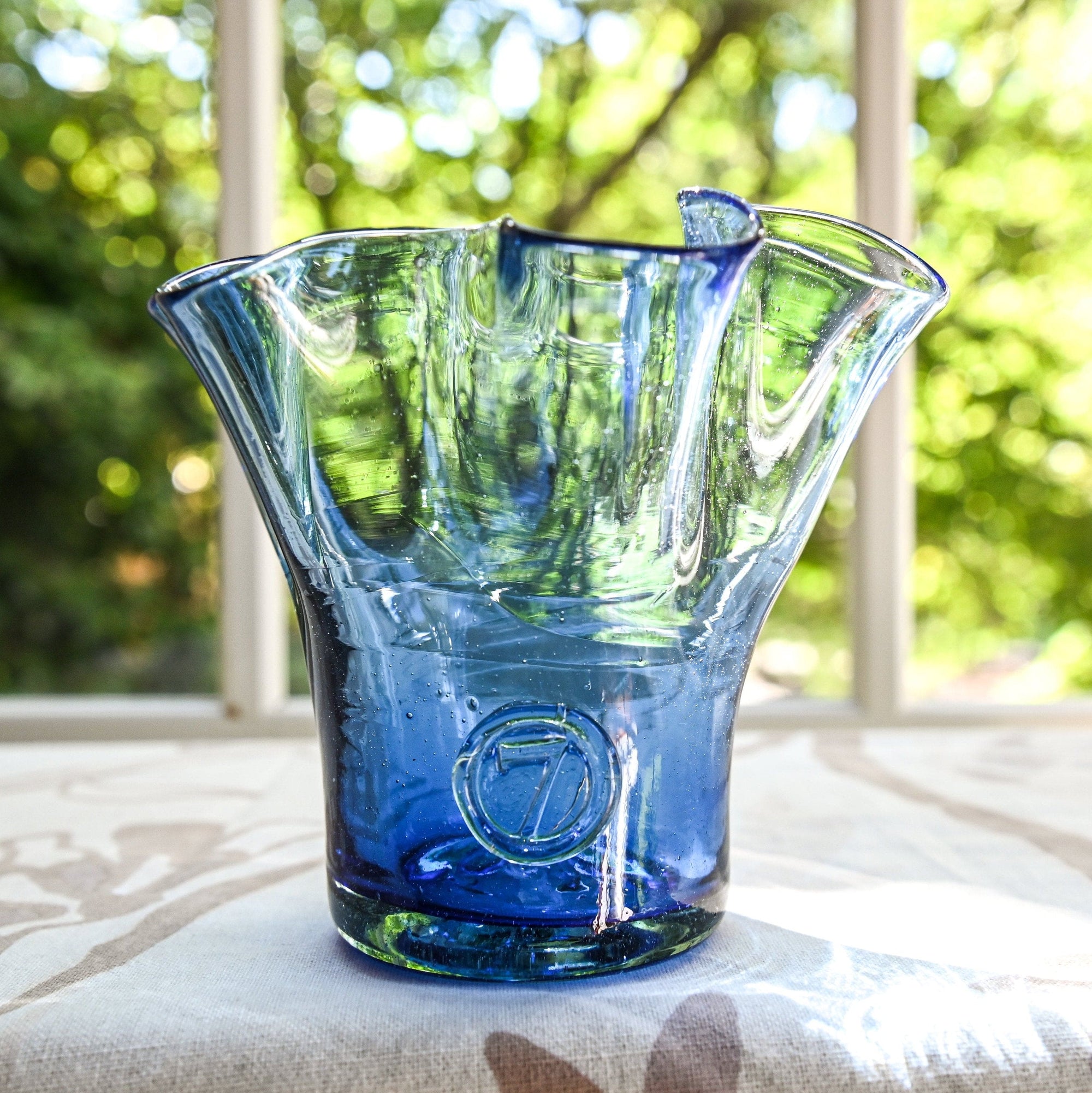 A handcrafted blue glass vase with a lettuce leaf pattern, placed on a table with a blurred background of greenery.