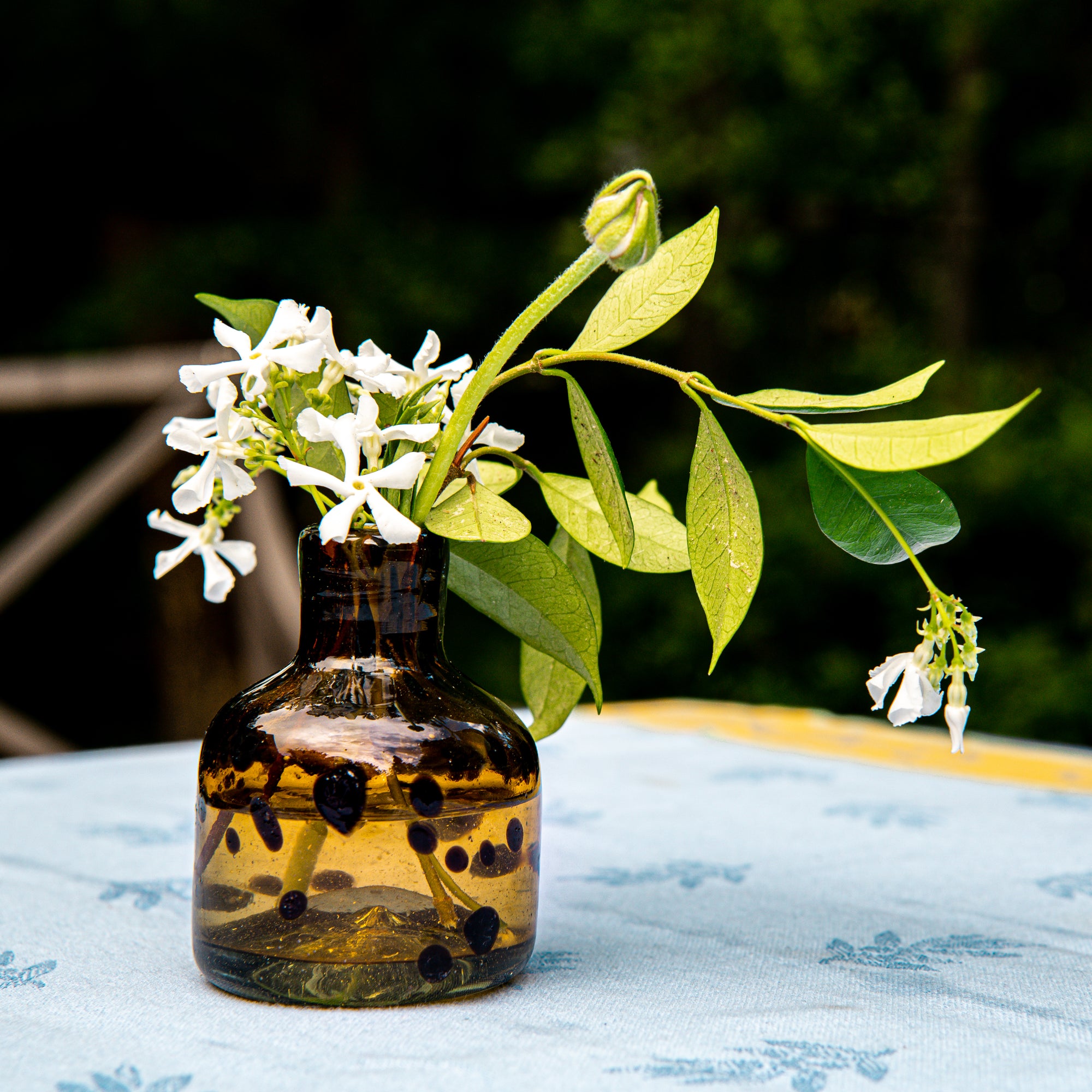 Small brown glass vase with white flowers and green leaves on a light blue tablecloth.