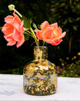 Decorative glass bottle with gold leaf design holding pink flowers on a table outdoors.