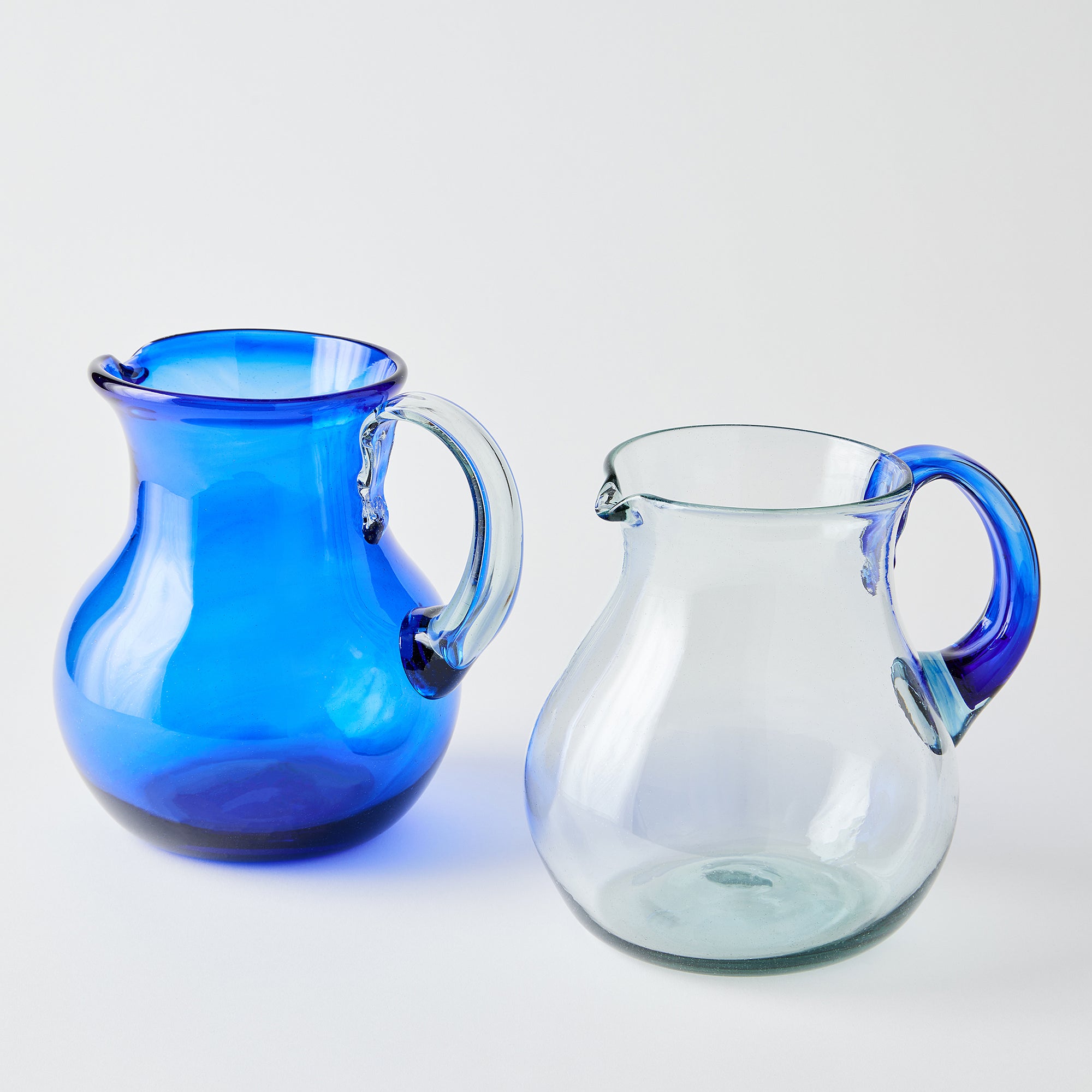 Two glass pitchers, one blue with a clear handle and one clear with a blue handle, on a white background