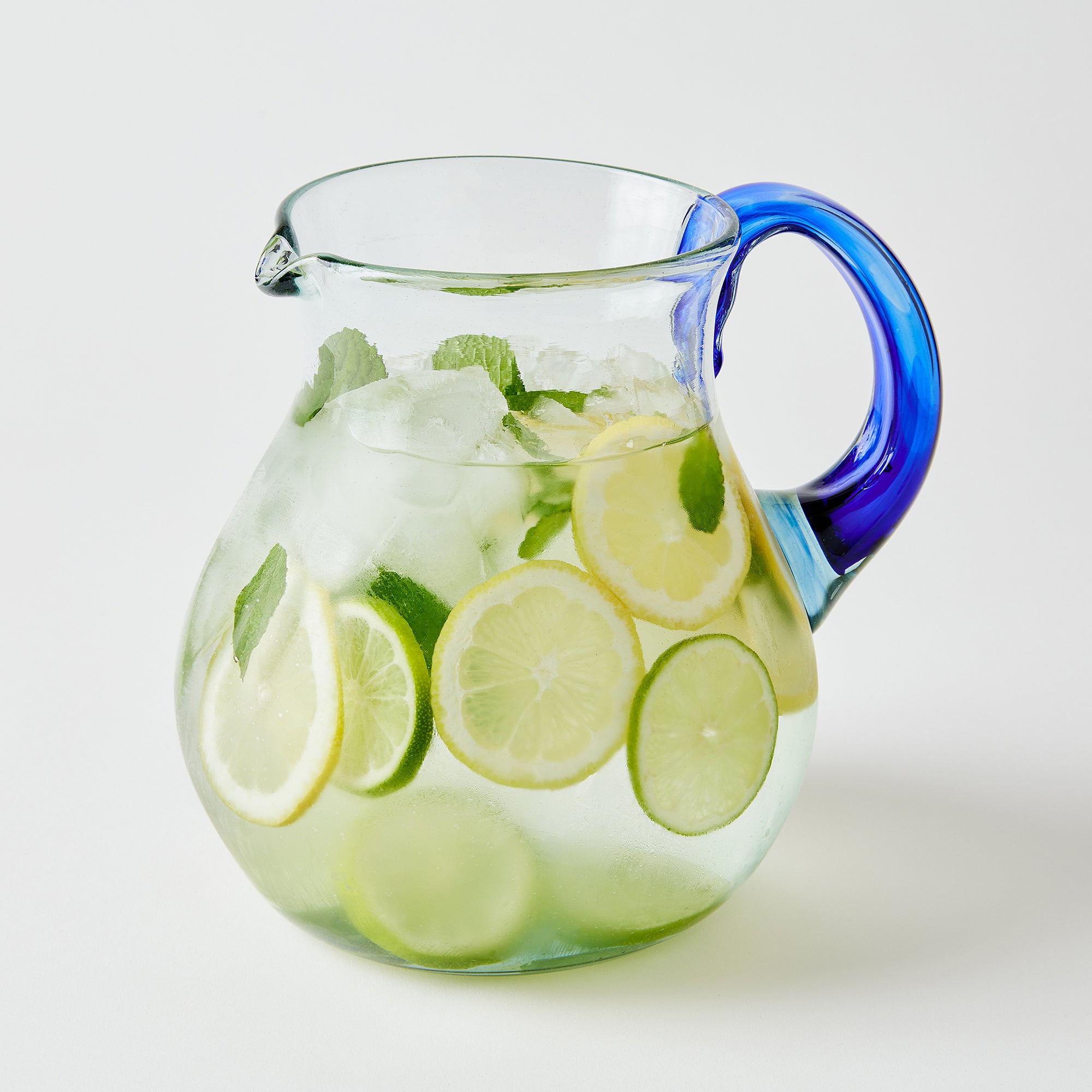 Clear glass pitcher with blue handle filled with lemon and lime slices infused drink and ice on a white background