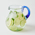 Clear glass pitcher with blue handle filled with lemon and lime slices infused drink and ice on a white background
