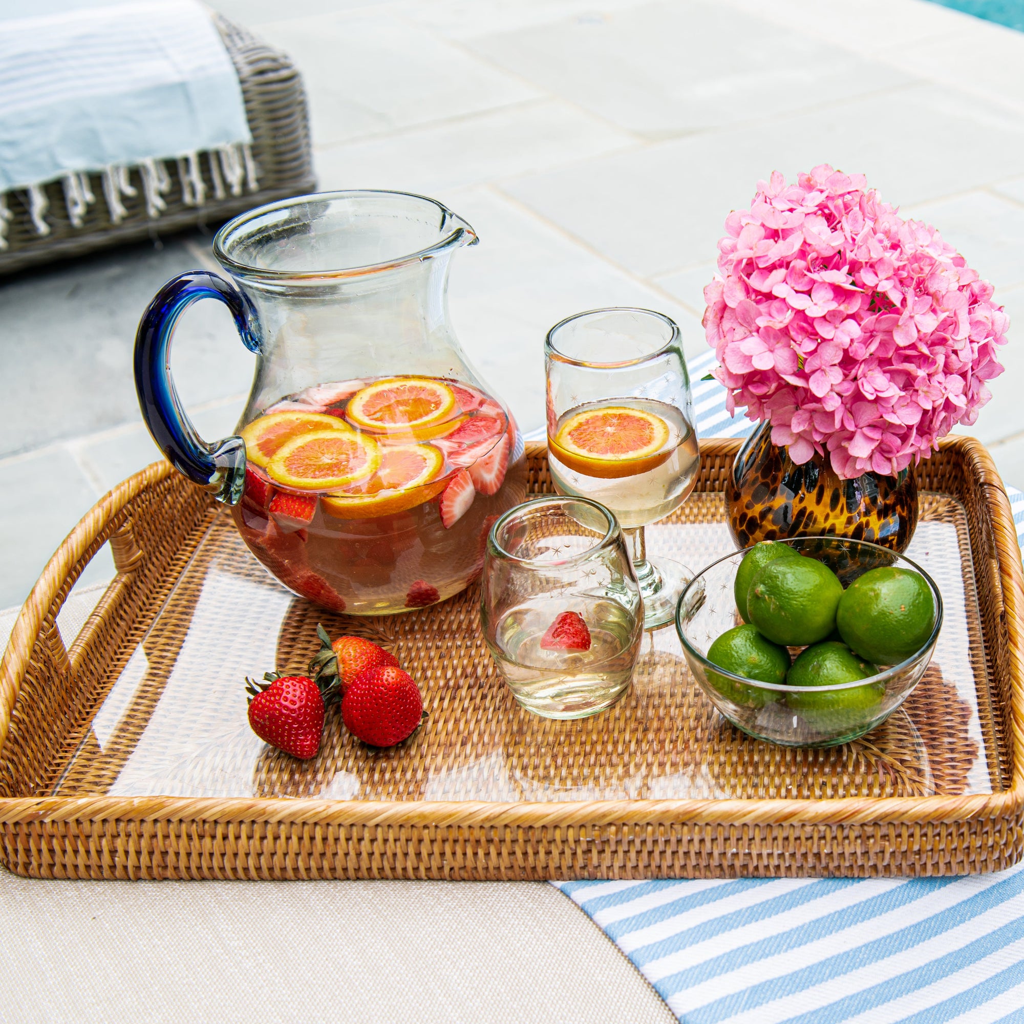 Tray with a pitcher of fruit-infused drink, glasses, vases, bowls, and fruits by a poolside.