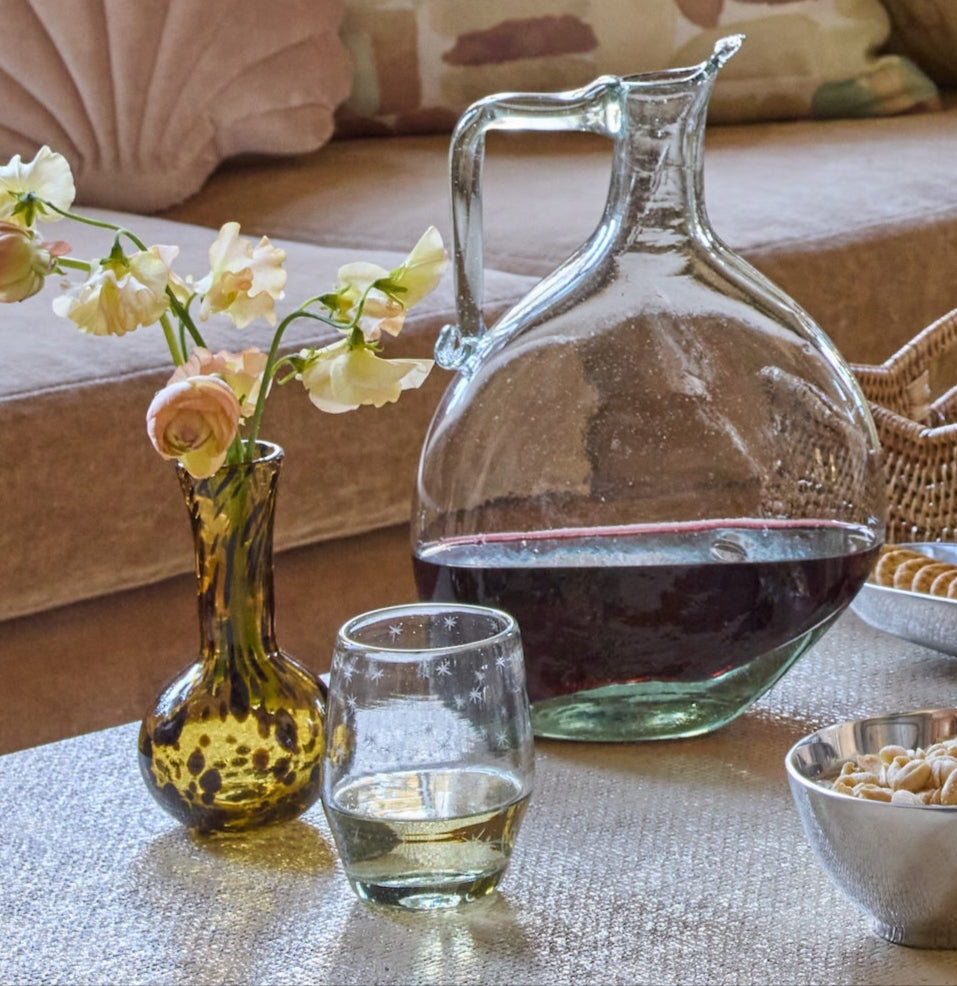 Glass decanter with red wine, glass of white wine, small vase with flowers, and bowl of snacks on a table.